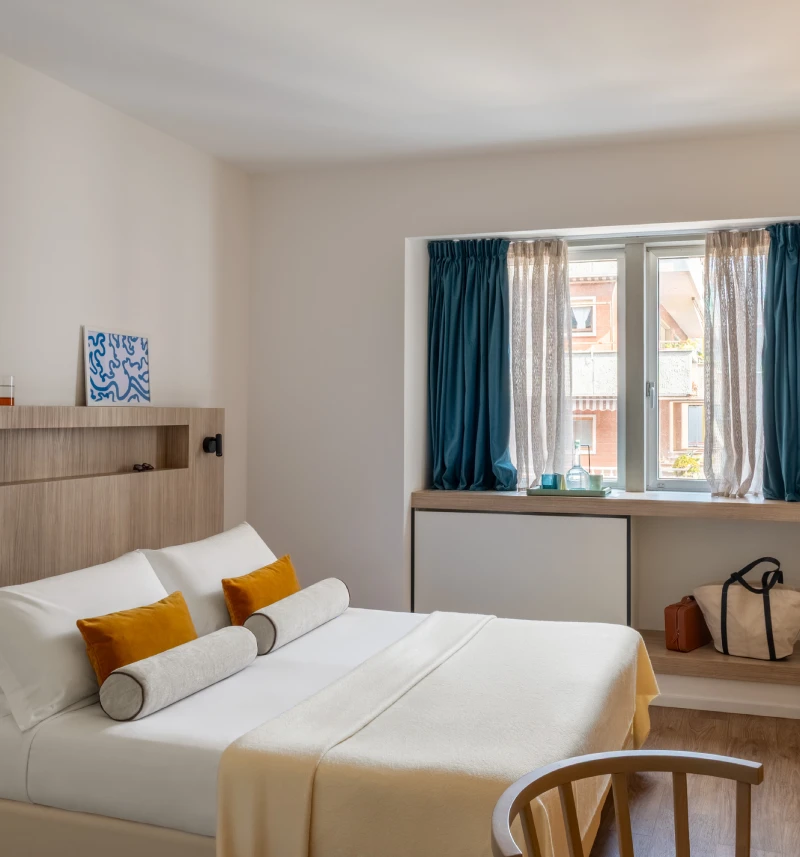 Bright double room with white bed, mustard cushions and window with blue curtains - Aparthotel Colombo, BZAR hotels in Rome