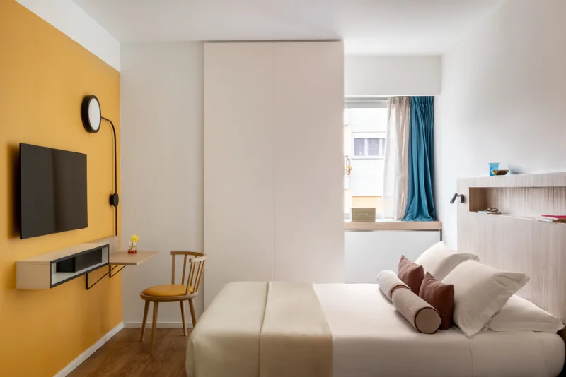 Double bedroom with ochre accent wall, flat-screen TV, work desk and natural light - Aparthotel Colombo, BZAR hotels in Rome
